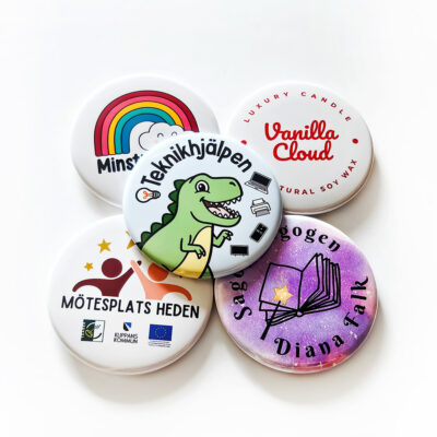 Fem runda pins och reklammagneter med olika logotyper och illustrationer, bland annat en regnbåge och en dinosaurie.